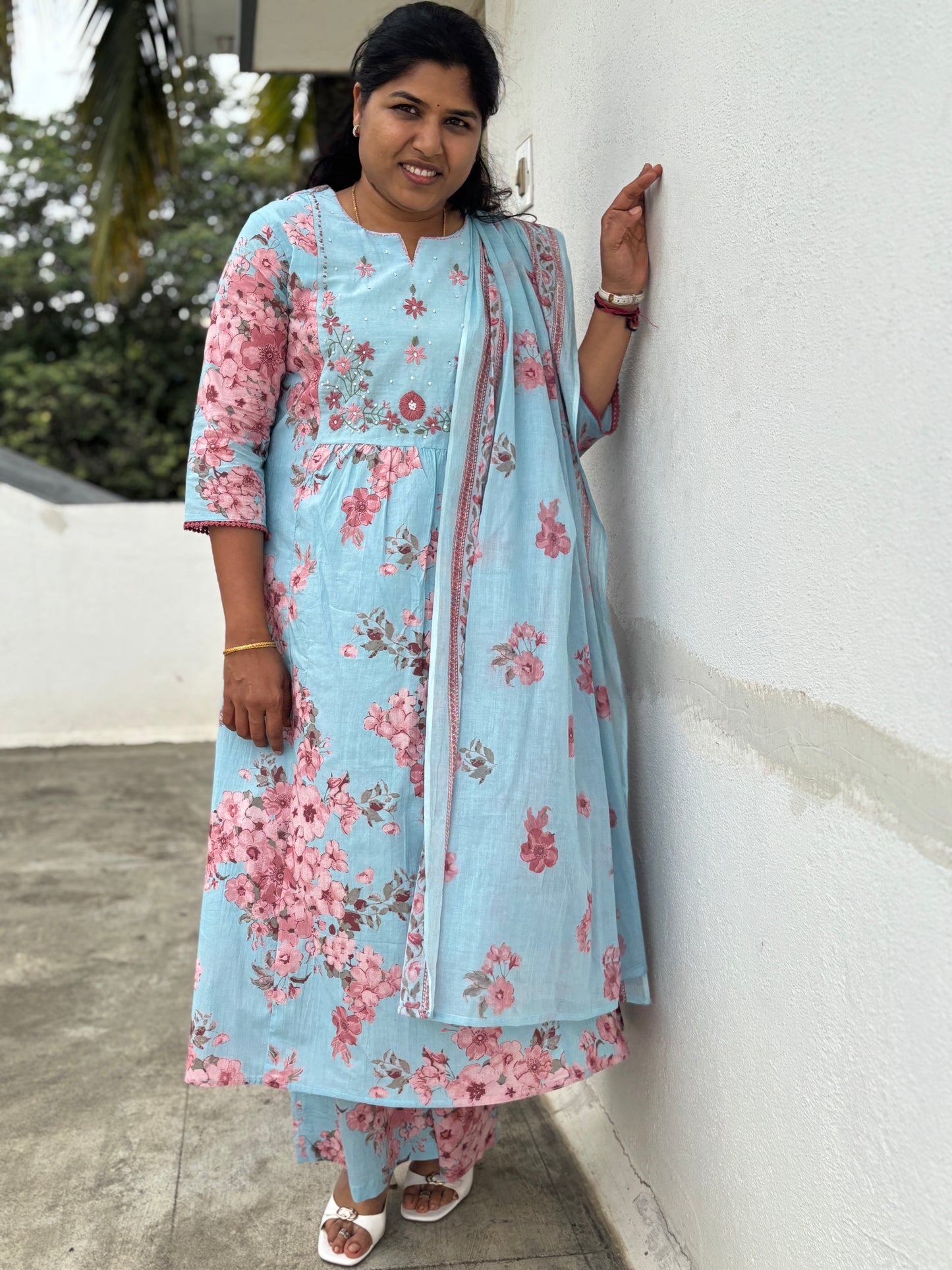 Woman in a light blue floral outfit leaning against a white wall.