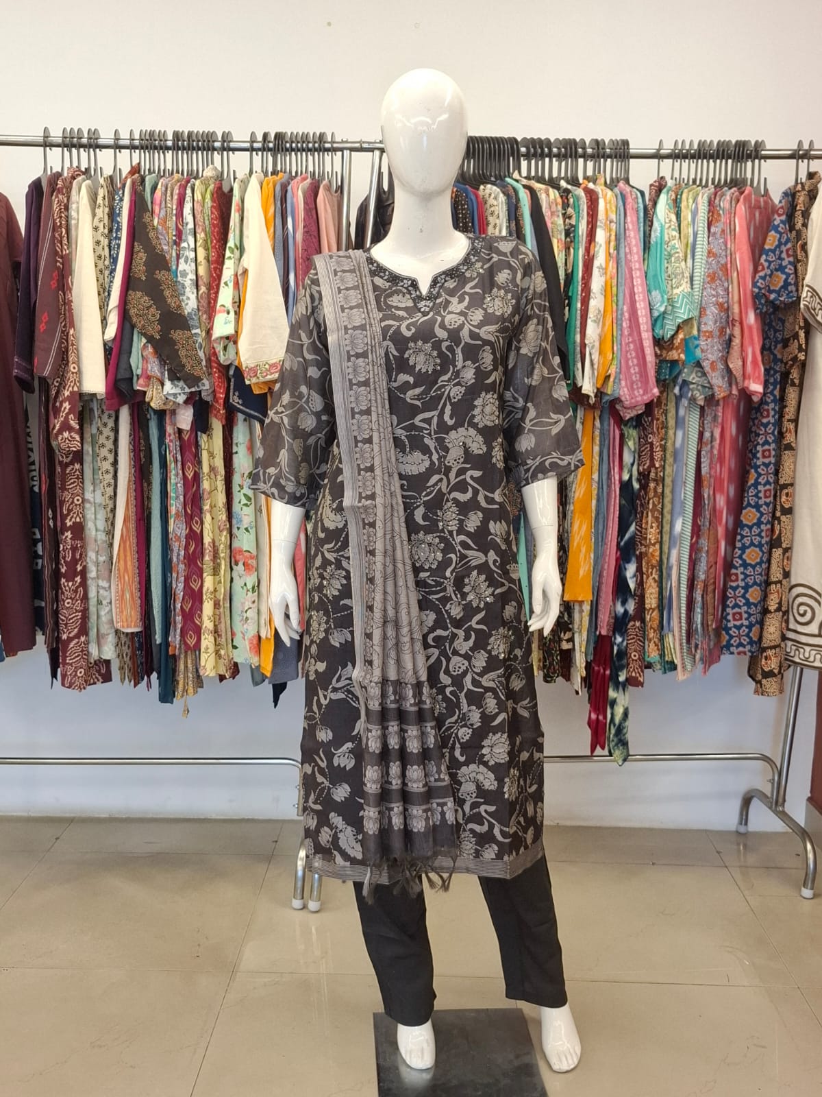 Mannequin wearing a black floral dress with a patterned shawl in front of a rack of colorful clothing.