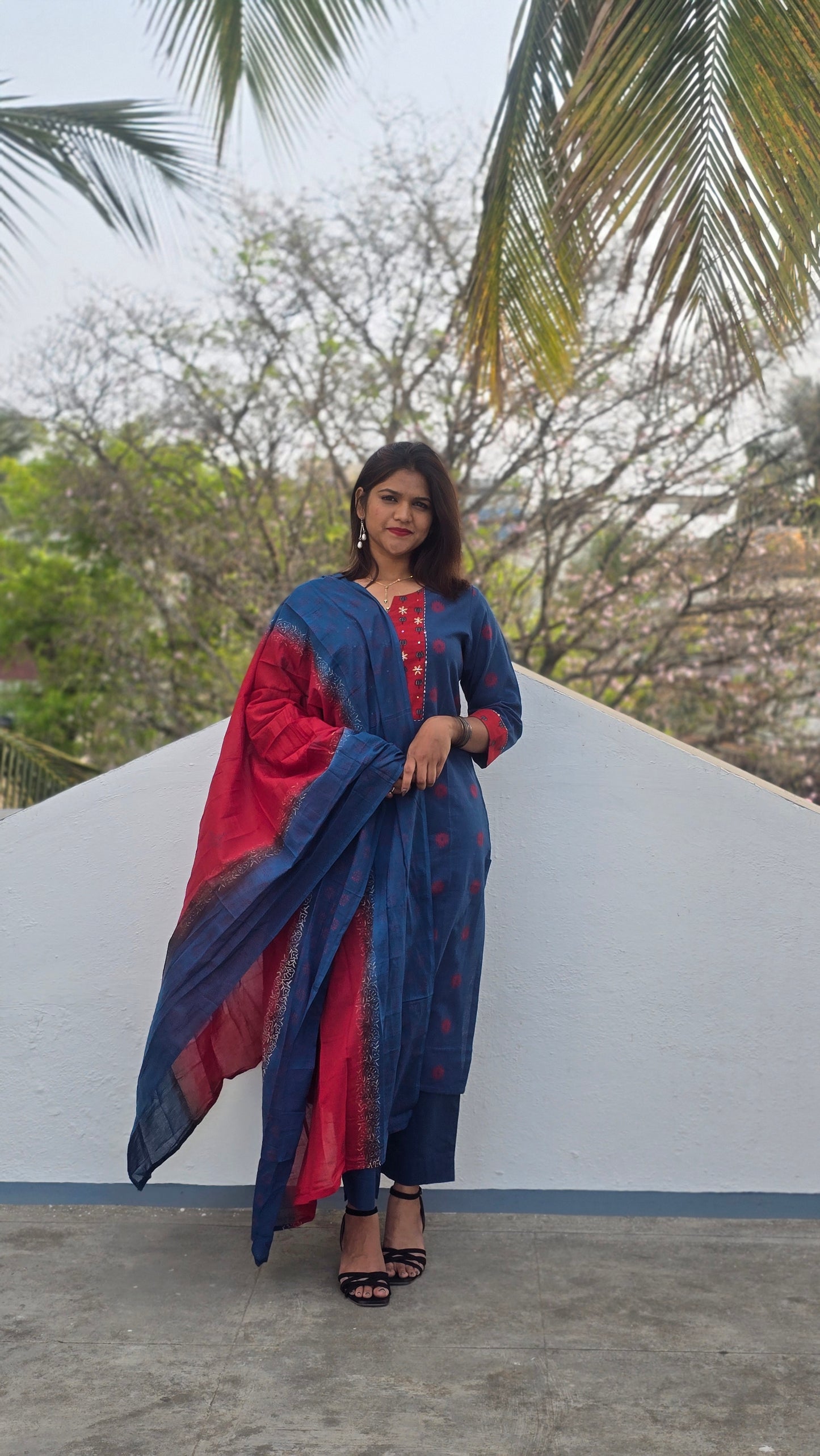 Woman in a blue and red traditional outfit standing against a white wall with palm trees in the background.