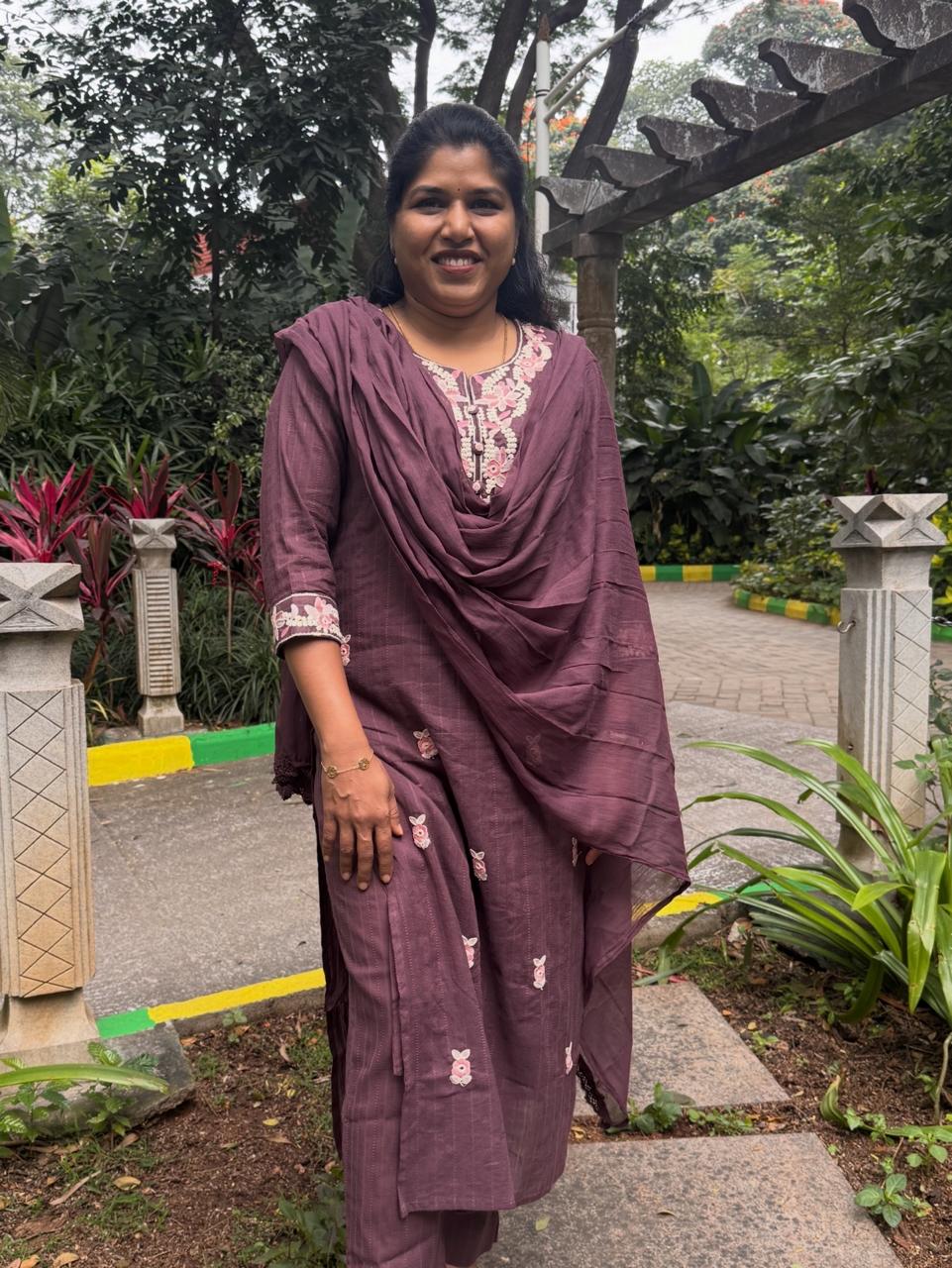 Woman in a purple traditional outfit standing outdoors with greenery in the background