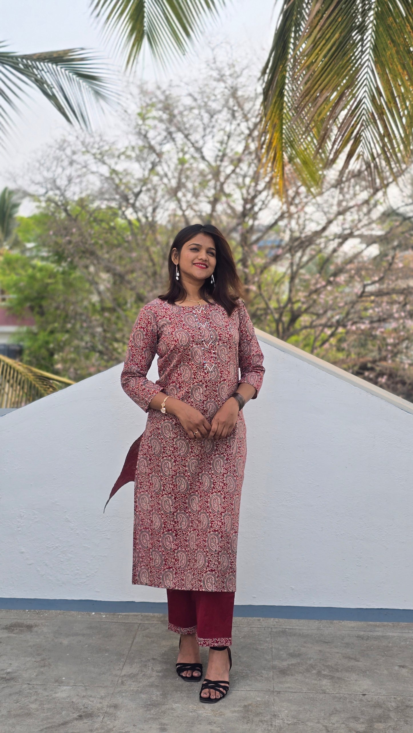 Woman in a patterned dress standing outdoors with trees and a white wall in the background