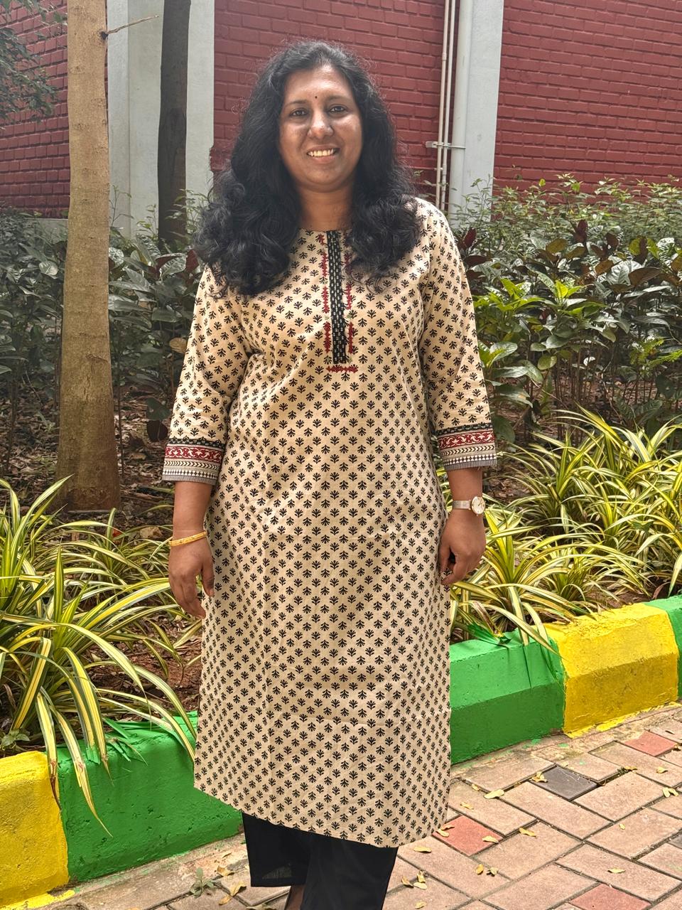 Woman in a patterned kurta standing outdoors with plants and a building in the background
