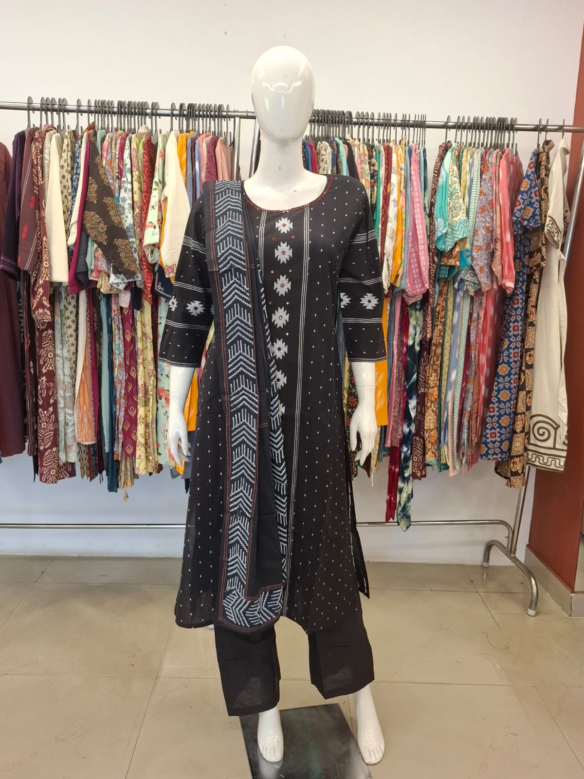 Mannequin wearing a black patterned outfit with a colorful clothing rack in the background.