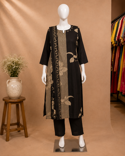A black three-piece kurta set displayed on a white mannequin, featuring a black tunic with beige floral patterns and a matching black-and-beige border drape, paired with black pants. The mannequin is set against a warm beige studio backdrop with a small wooden stool and vase on the left, and a clothing rack visible on the right.
