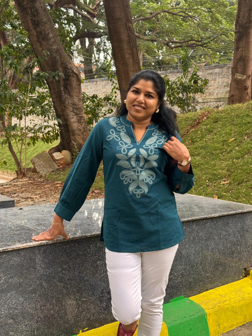 Woman in a teal embroidered blouse and white pants standing outdoors with trees and grass in the background.