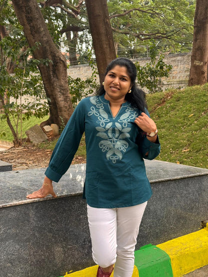 Woman in a teal embroidered blouse and white pants standing outdoors with trees and grass in the background.