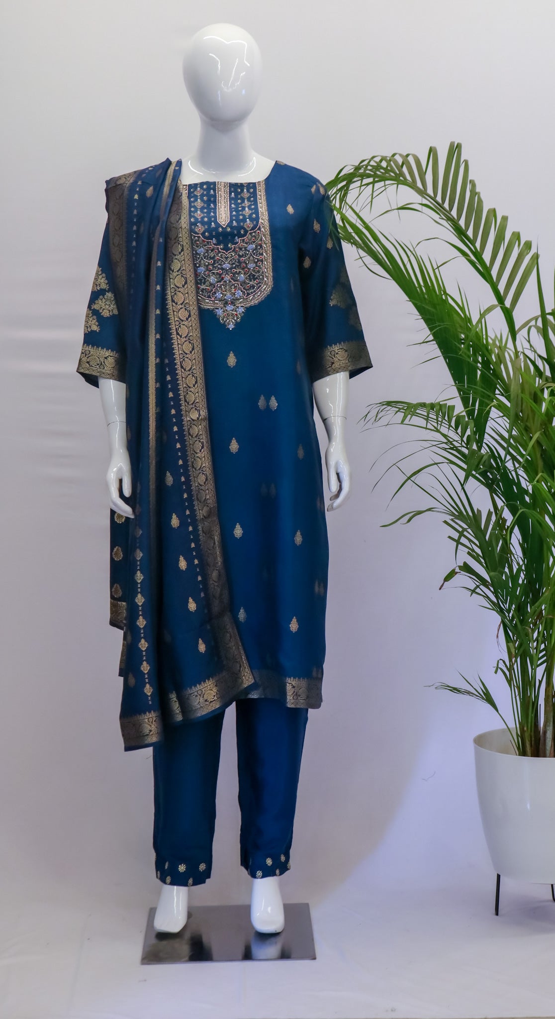 A navy blue silk dupatta set displayed on a mannequin, consisting of a kurta with pockets, a pant, and a dupatta with zari work, arranged against a backdrop of a potted plant and a white panel.