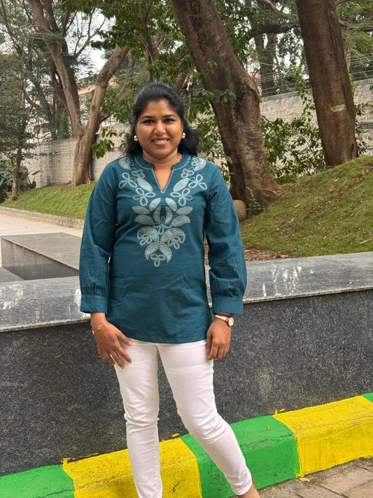 Woman wearing a teal embroidered blouse and white pants standing outdoors.