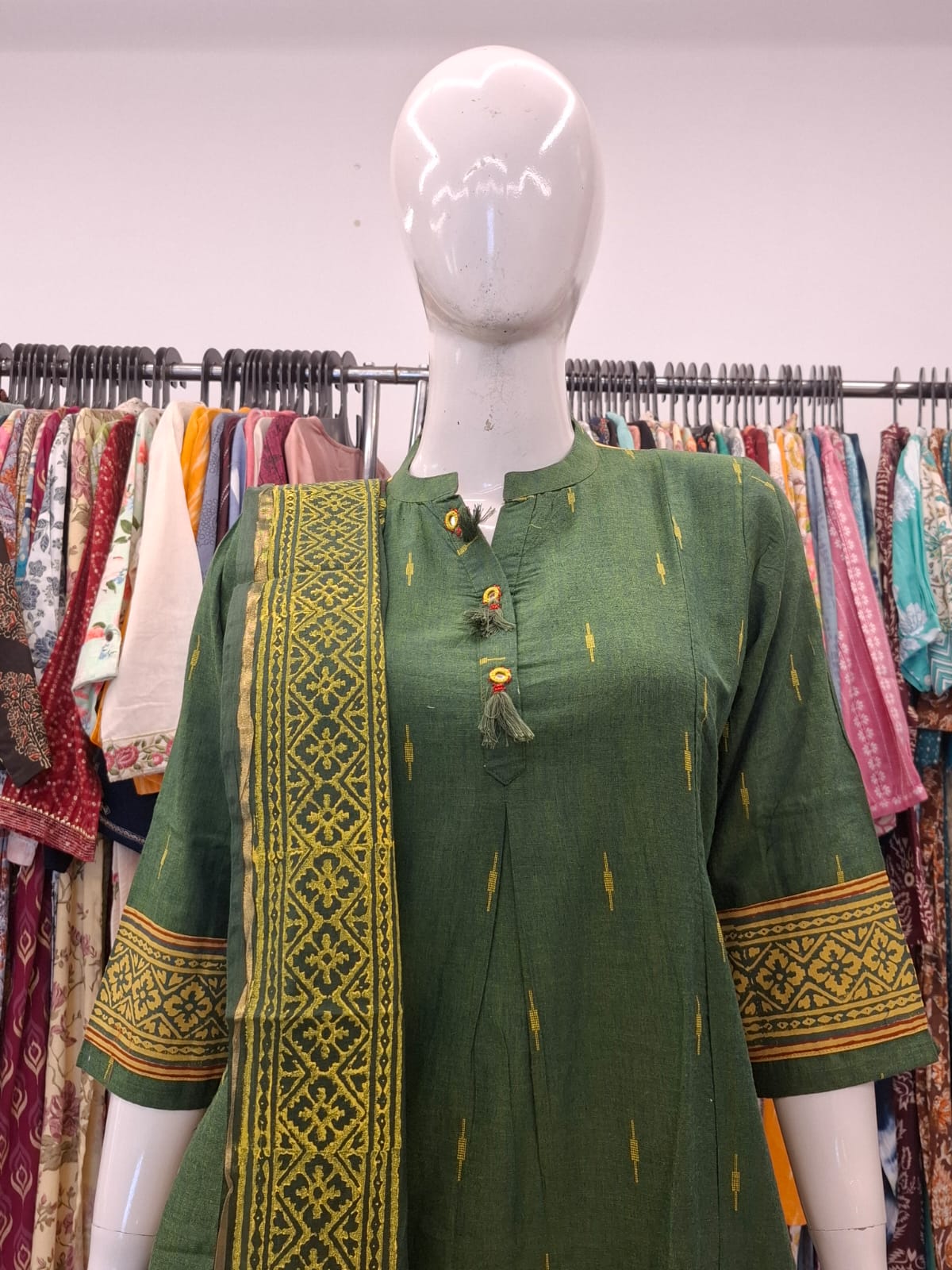 Green embroidered kurta on a mannequin with a colorful background of clothes.