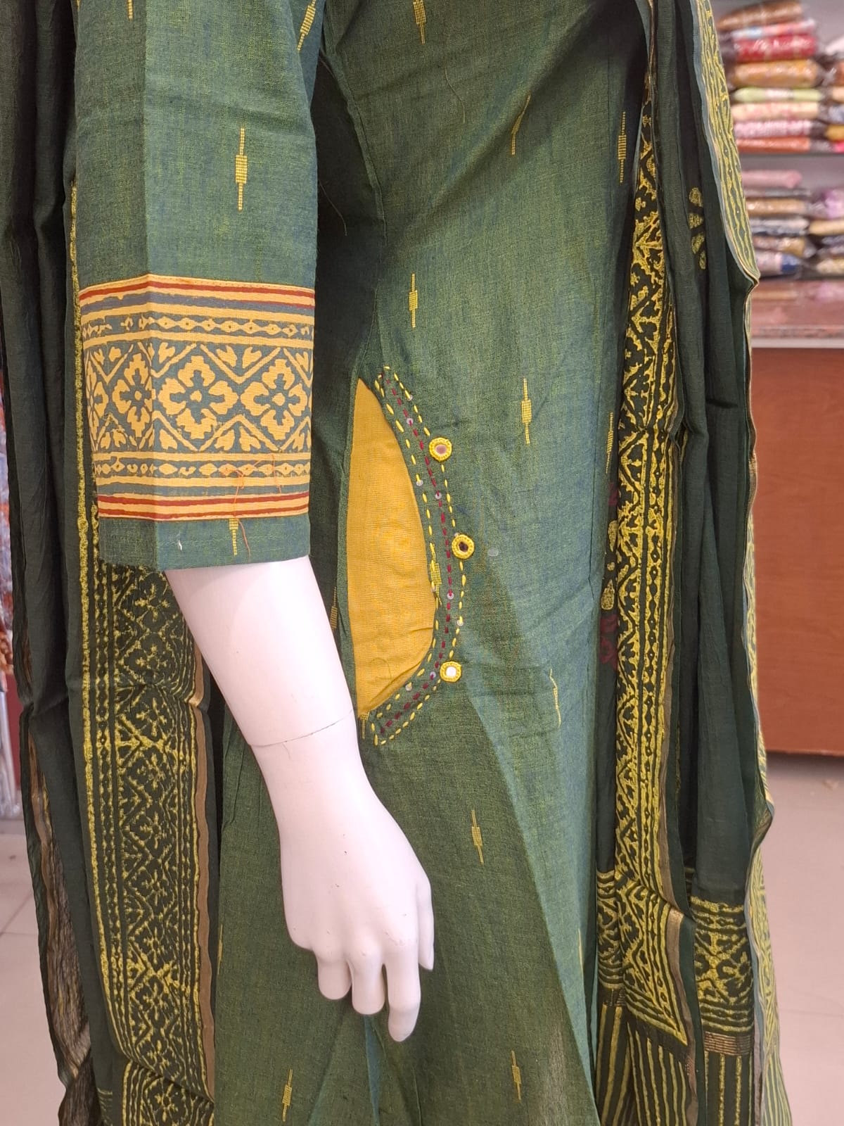Green traditional outfit with yellow patterns on a mannequin in a store setting.
