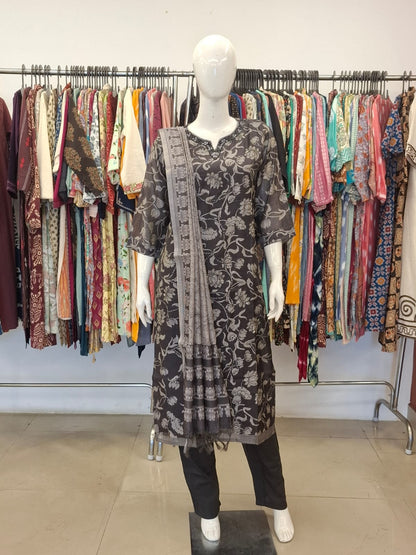 Mannequin wearing a black floral dress with a patterned shawl in front of a rack of colorful clothing.