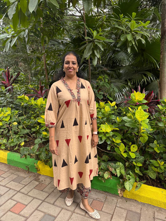 Woman in a beige dress with black and red triangle patterns standing in front of green foliage.
