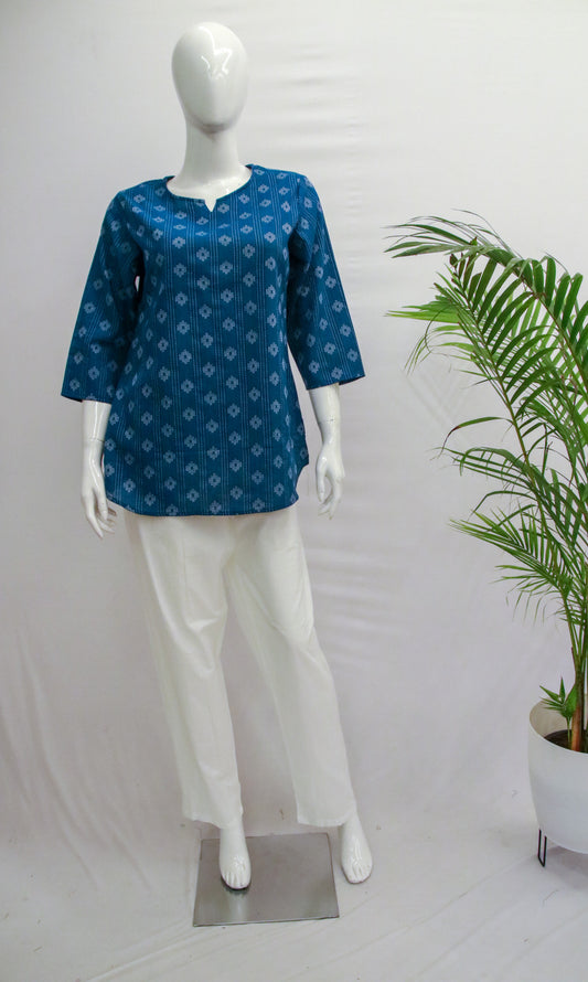 Mannequin wearing a blue patterned top and white pants with a plant in the background.
