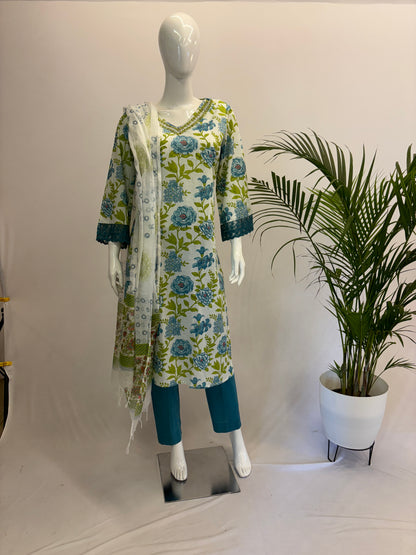A mannequin displaying a white, blue, and green floral dupatta set with a V-neck kurta and pants. There is a potted plant beside the mannequin.