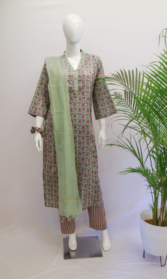 A mannequin displaying a green floral dupatta set with a kurta and pant, standing in front of a white backdrop with a potted plant to the side.