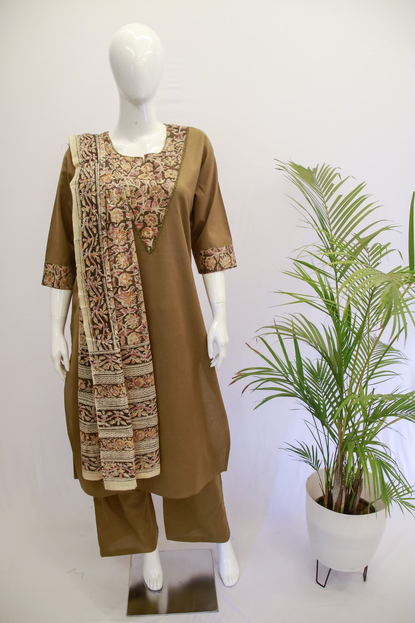 A mannequin displaying a green and beige traditional Indian cotton Mehndi dupatta set with embroidery, alongside a potted plant.