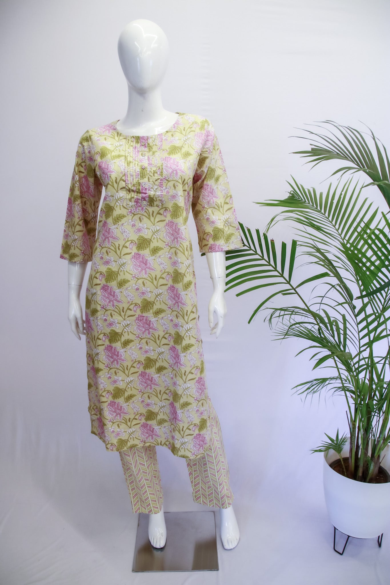A mannequin displaying a green top and pant set with floral prints, standing in a retail setting with a potted plant beside it.
