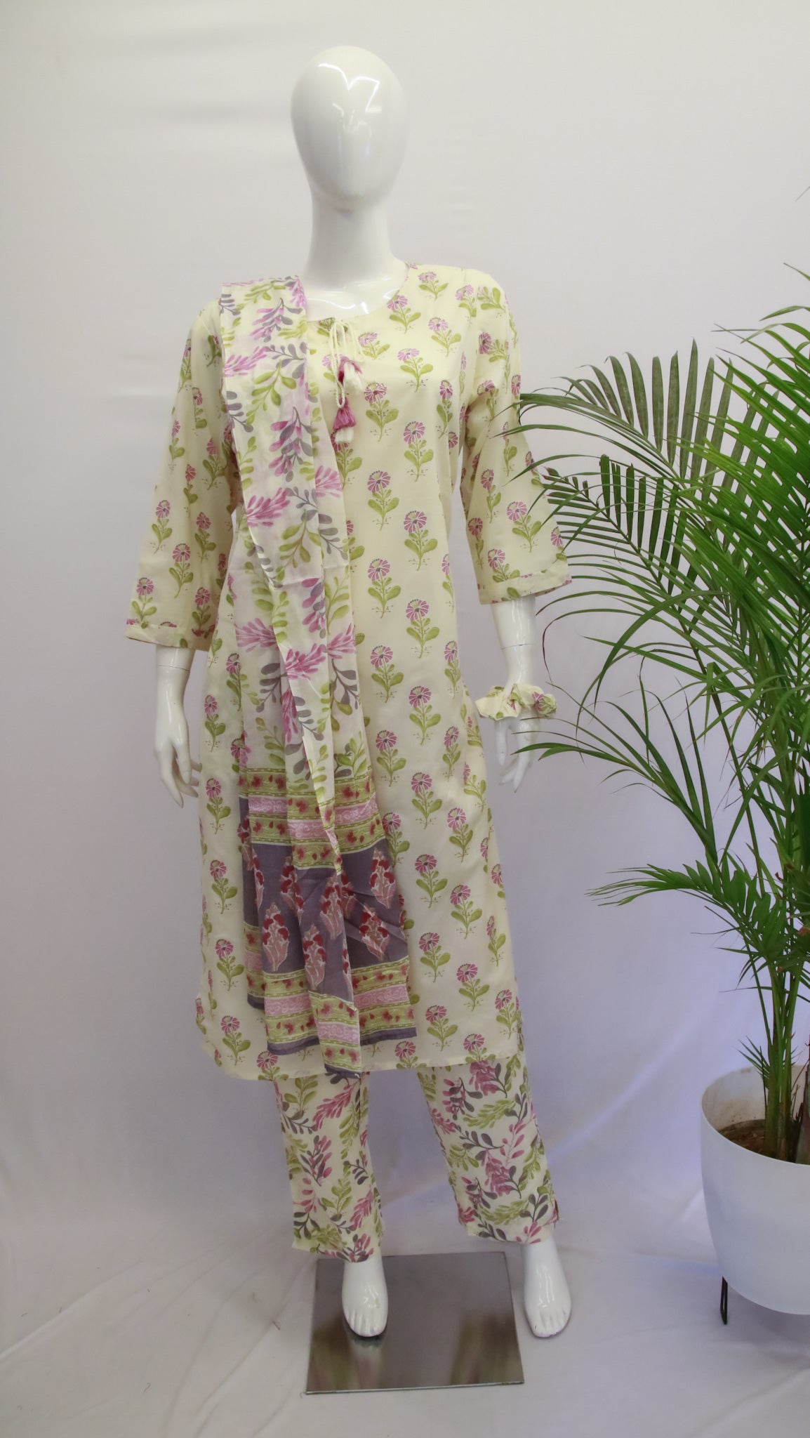 A mannequin displaying a white floral dupatta set with a kurta, pant, and dupatta, standing in a room with a potted plant in the background.