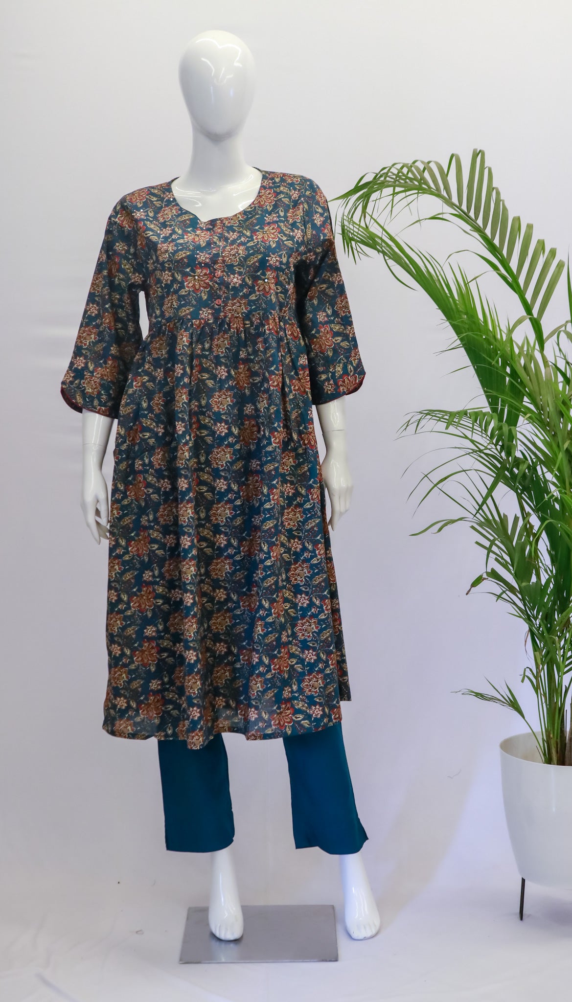 A mannequin displaying a green nyra top and pant set with a floral print, standing in front of a white background with a potted plant to the side.
