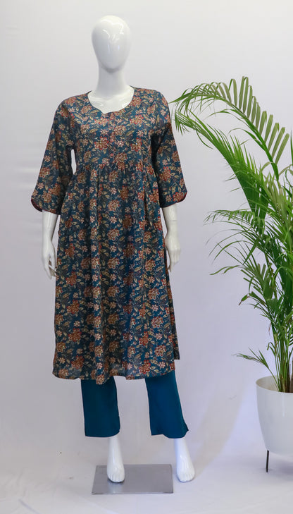 A mannequin displaying a green nyra top and pant set with a floral print, standing in front of a white background with a potted plant to the side.