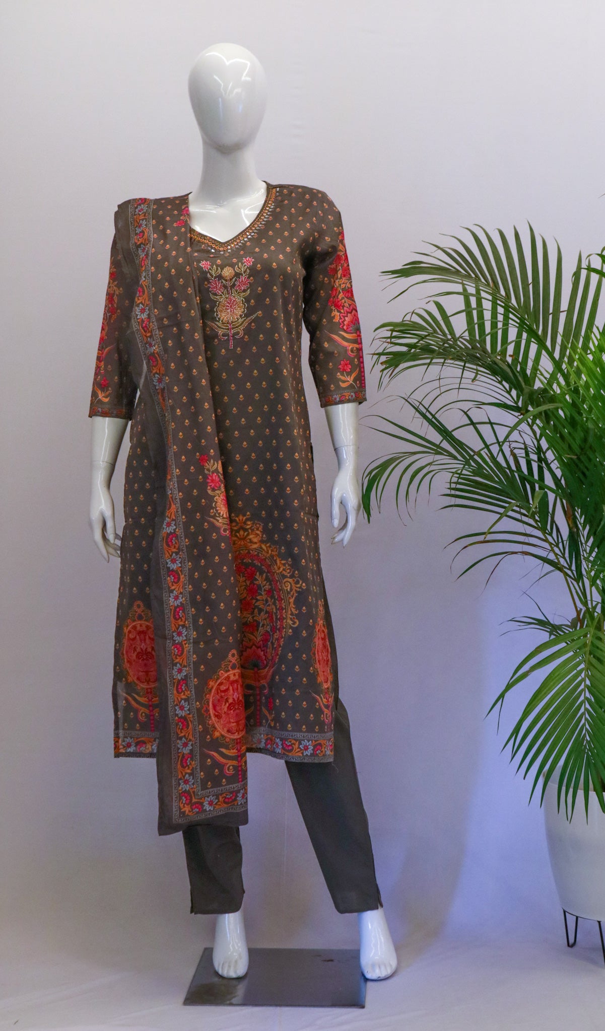 A mannequin displaying a grey cotton dupatta set with orange and dark gray prints, including a kurta, pant, and dupatta, standing in a room with a potted plant in the background.