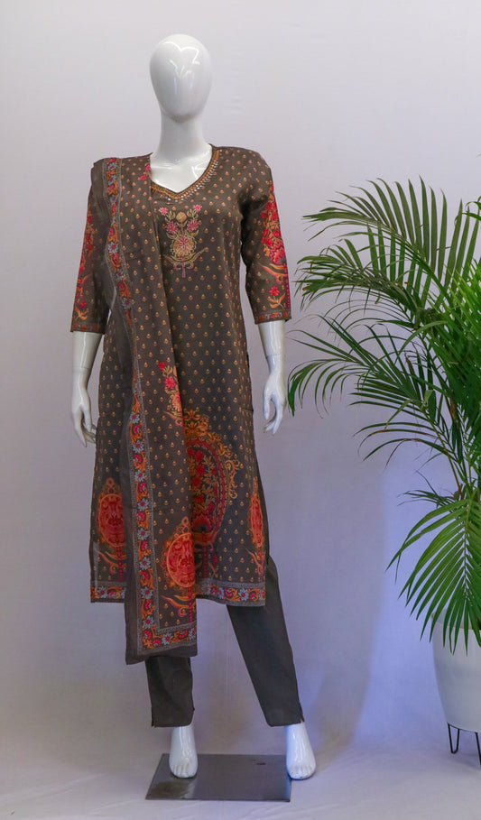 A mannequin displaying a grey cotton dupatta set with orange and dark gray prints, including a kurta, pant, and dupatta, standing in a room with a potted plant in the background.