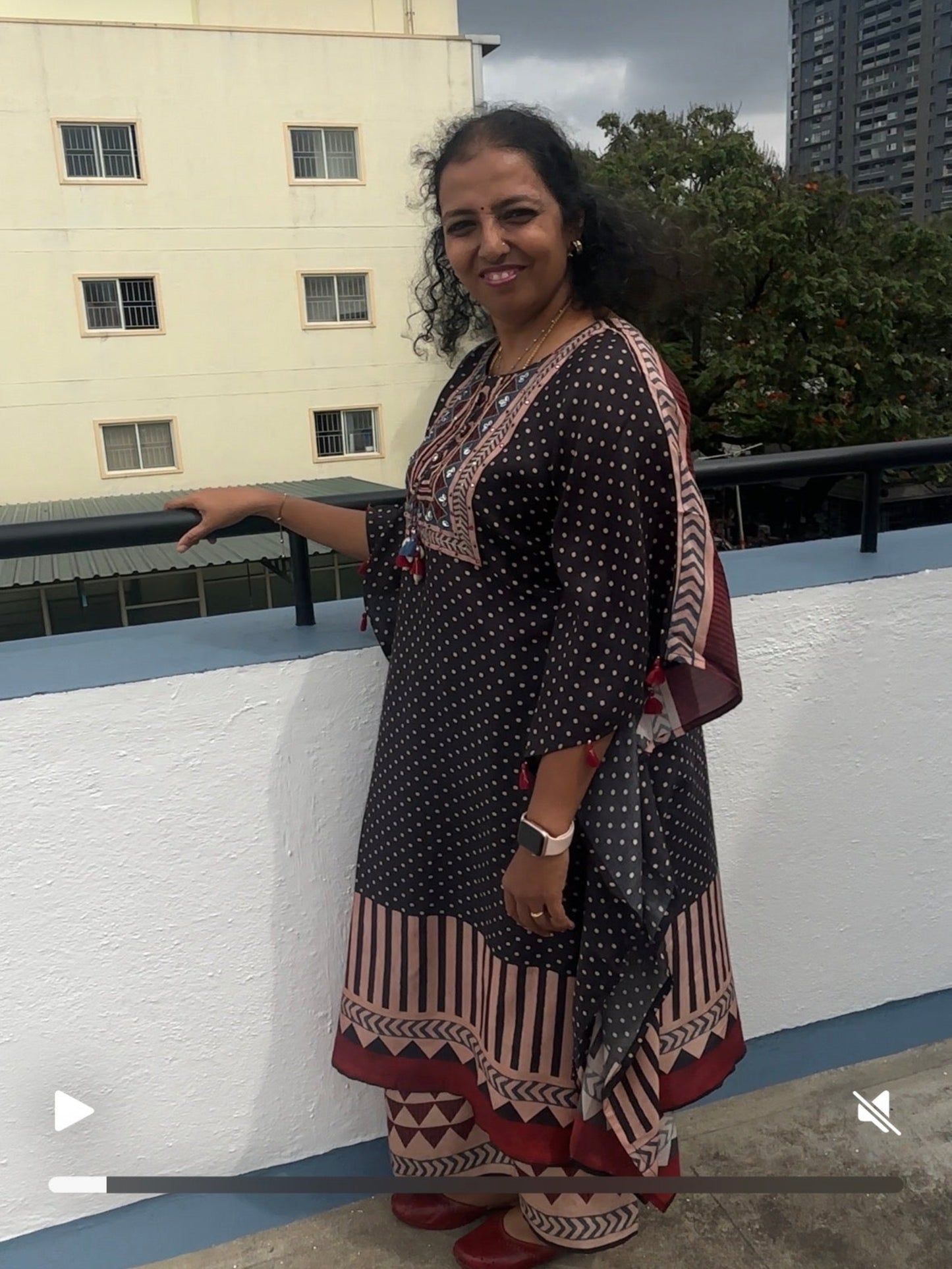 Woman in a patterned dress standing on a rooftop with buildings and trees in the background