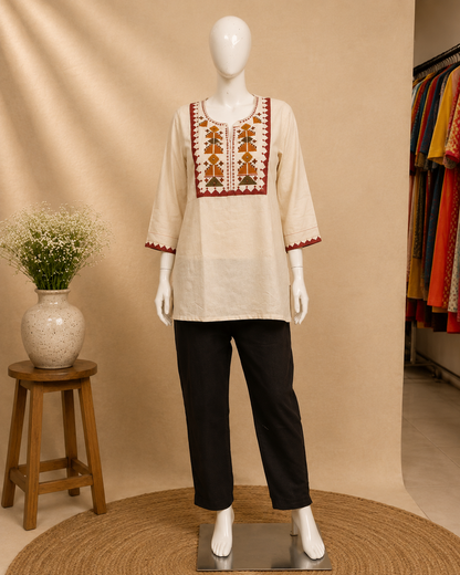 A cream-colored cotton kurta with red and gold geometric embroidery on the chest and sleeve cuffs, styled with black trousers on a white mannequin. The mannequin is set against a warm beige backdrop with a small wooden stool and a vase of flowers on the left, and a rack of colorful garments on the right.