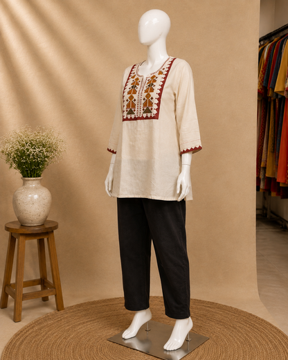 A cream-colored linen tunic top with brown and red geometric embroidery on the chest and sleeve cuffs, styled on a female mannequin with black cropped trousers, set against a warm beige studio backdrop with a small wooden stool and vase of flowers.