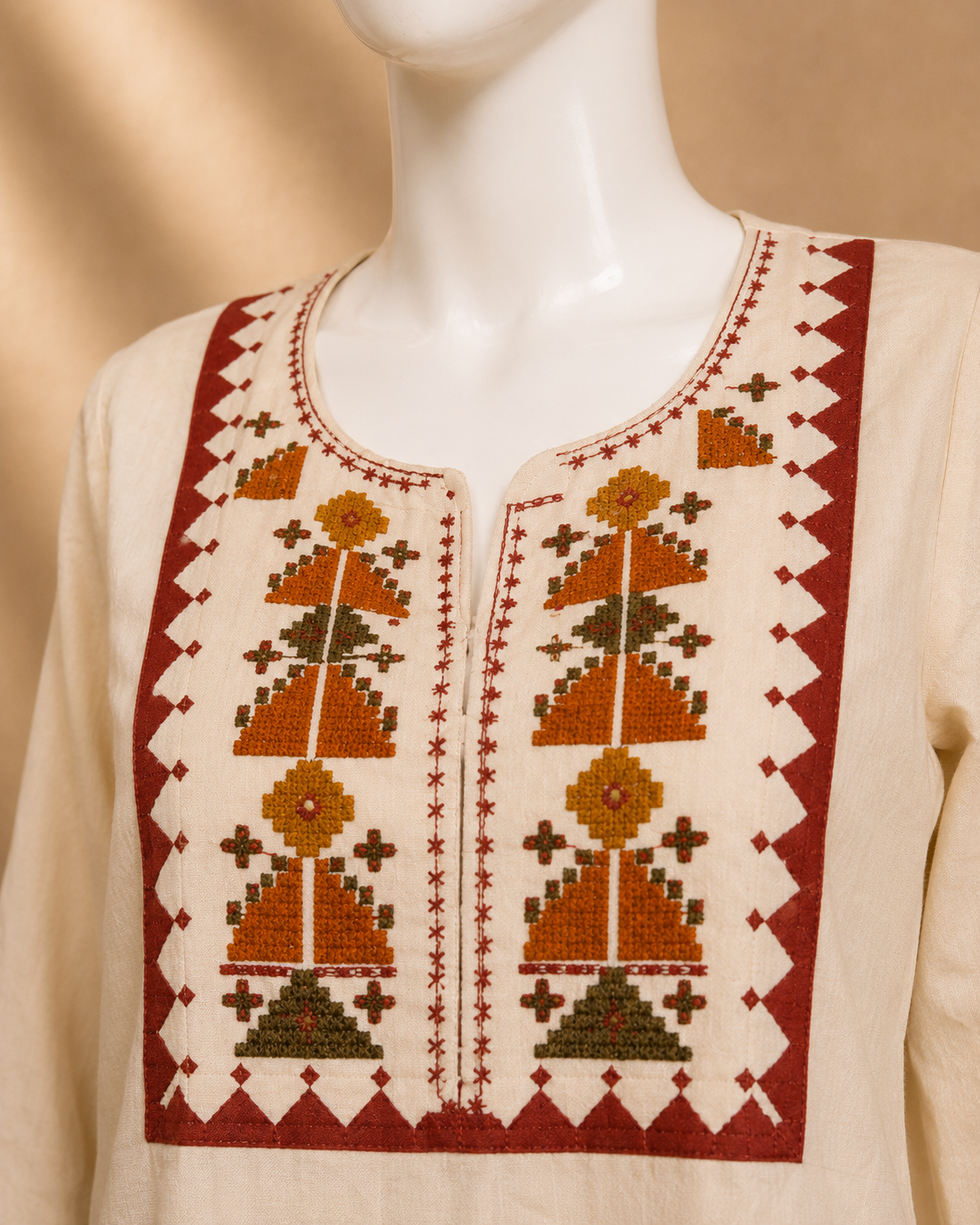 A cream-colored embroidered tunic top displayed on a white mannequin torso. The garment features a wide red border framing a central panel with geometric and tree-like patterns in rust orange, olive green, and white.