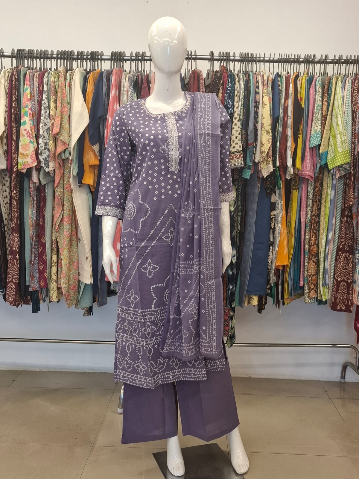Mannequin wearing a purple patterned dress in front of a clothing rack with various garments.