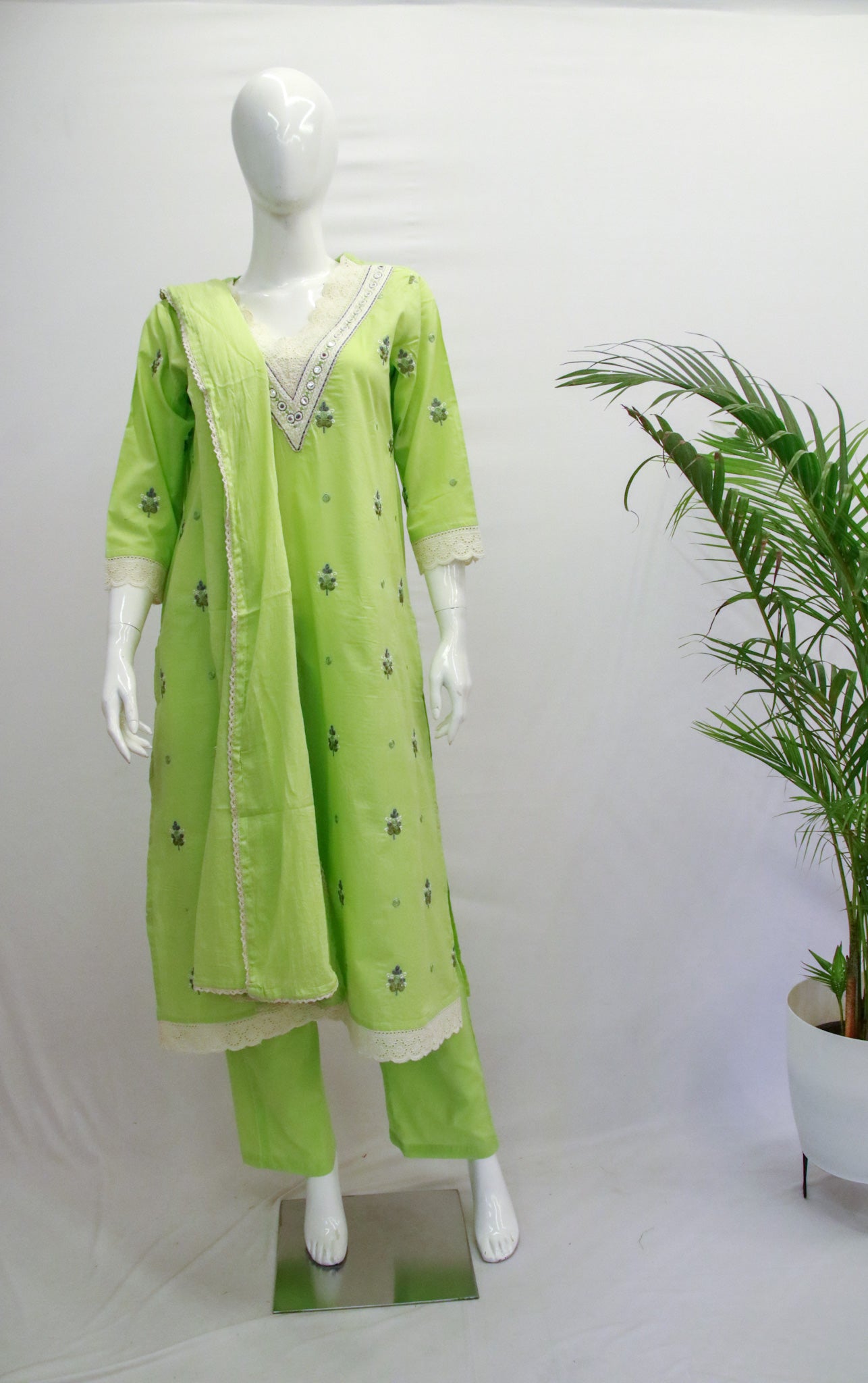 A mannequin displaying a green dupatta set with embroidery, consisting of a kurta, pant, and dupatta, against a white background with a potted plant beside it.