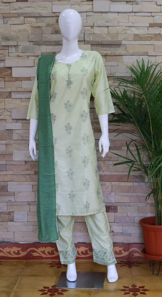 A traditional silk suit set in light green color with a crepe dupatta, displayed on a mannequin against a backdrop of stone walls and potted plants.