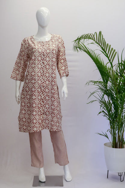 A mannequin displaying a pink and green floral muslin top and pants with a round neck and 3/4th sleeves, standing in a room with a potted plant in the background.