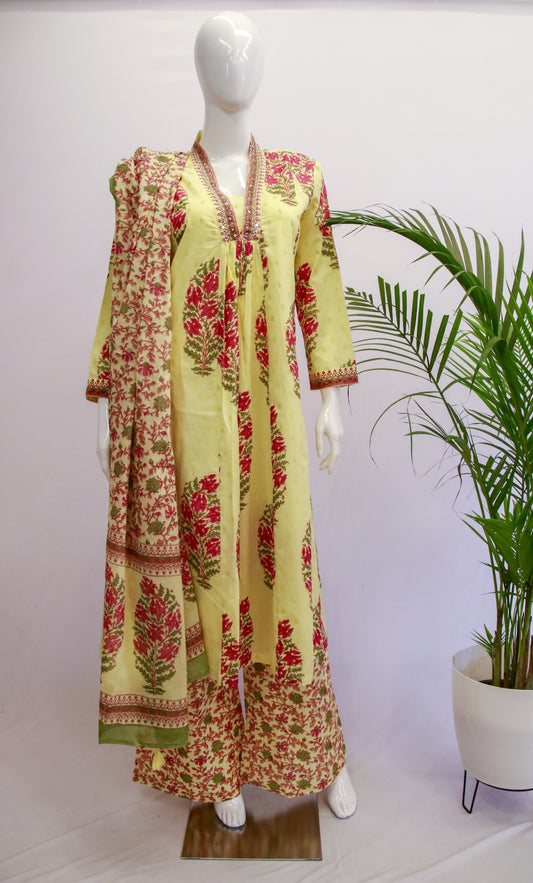 A mannequin displaying a yellow floral dupatta set with kurta and pants, standing against a white background with a potted plant beside it.