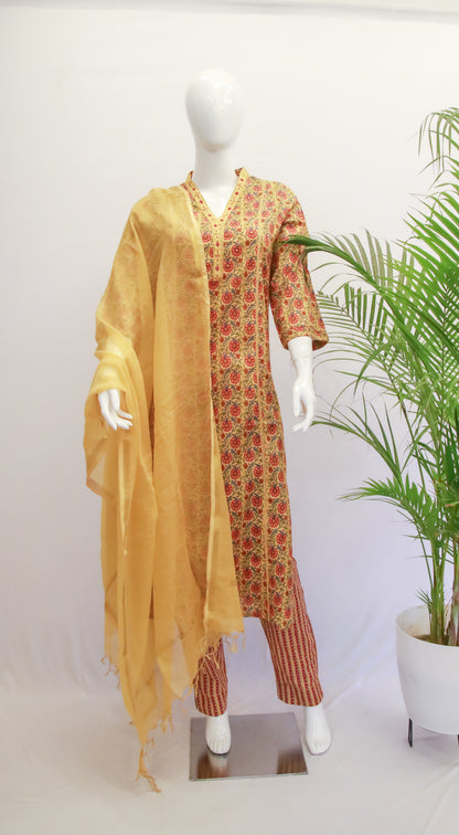 A mustard yellow cotton kurta set with floral dupatta and pants displayed on a mannequin, with a potted plant in the background.