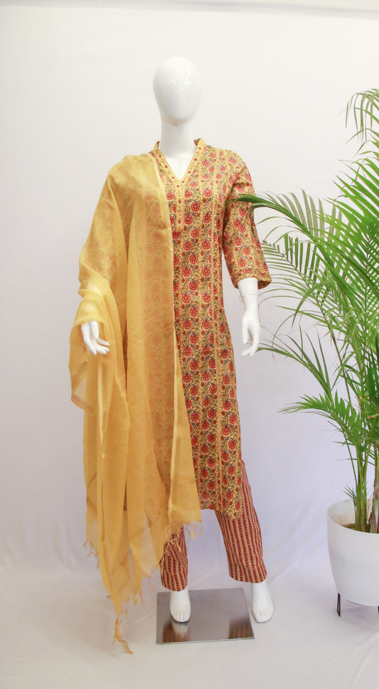 A mustard yellow cotton kurta set with floral dupatta and pants displayed on a mannequin, with a potted plant in the background.