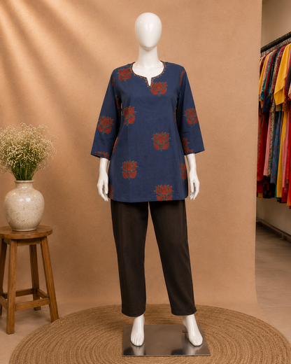 A mannequin wearing a navy blue three-quarter sleeve kurta with red floral motifs and black trousers, displayed against a warm beige studio backdrop with a small wooden stool and vase on the left.