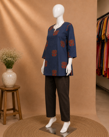 A mannequin wearing a navy blue tunic top with rust-red floral prints and dark brown wide-leg pants, displayed in a styled studio setting with a tan backdrop, woven rug, stool, and hanging garments in the background.