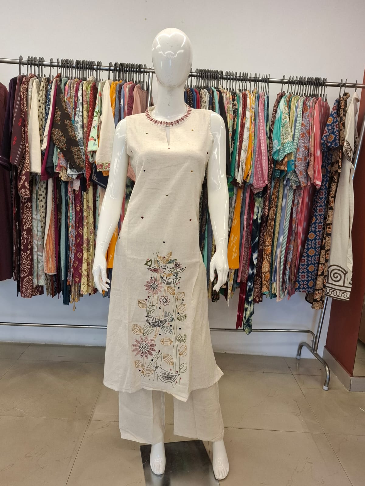 Mannequin wearing a white embroidered dress with a colorful clothing rack in the background.