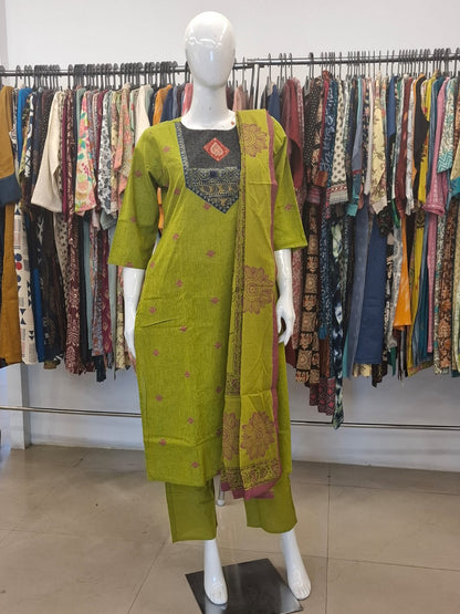 Mannequin wearing a green traditional outfit with a colorful background of hanging clothes.