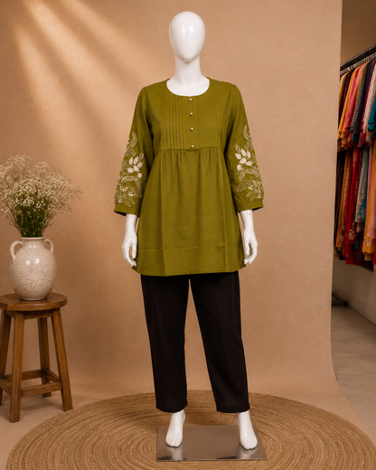 A mannequin wearing a loose olive green tunic top with white floral embroidery on the sleeves and black pants, displayed in a boutique setting against a warm beige backdrop.