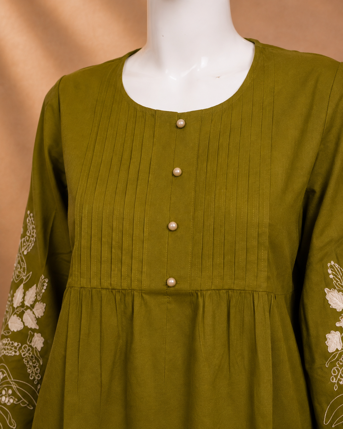 An olive green long-sleeve tunic top with vertical pleats on the bodice, a round neckline, decorative pearl-like buttons down the front, and white floral embroidery on the sleeves, displayed on a white mannequin torso.