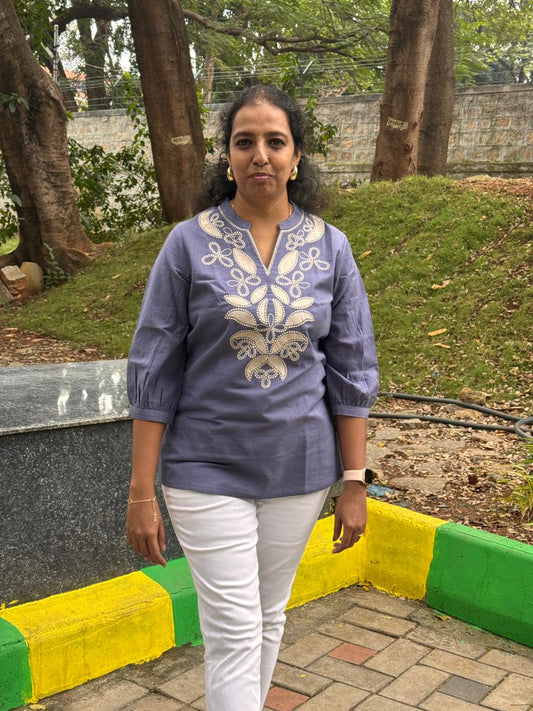Woman wearing a blue embroidered top and white pants standing outdoors with trees and a wall in the background.