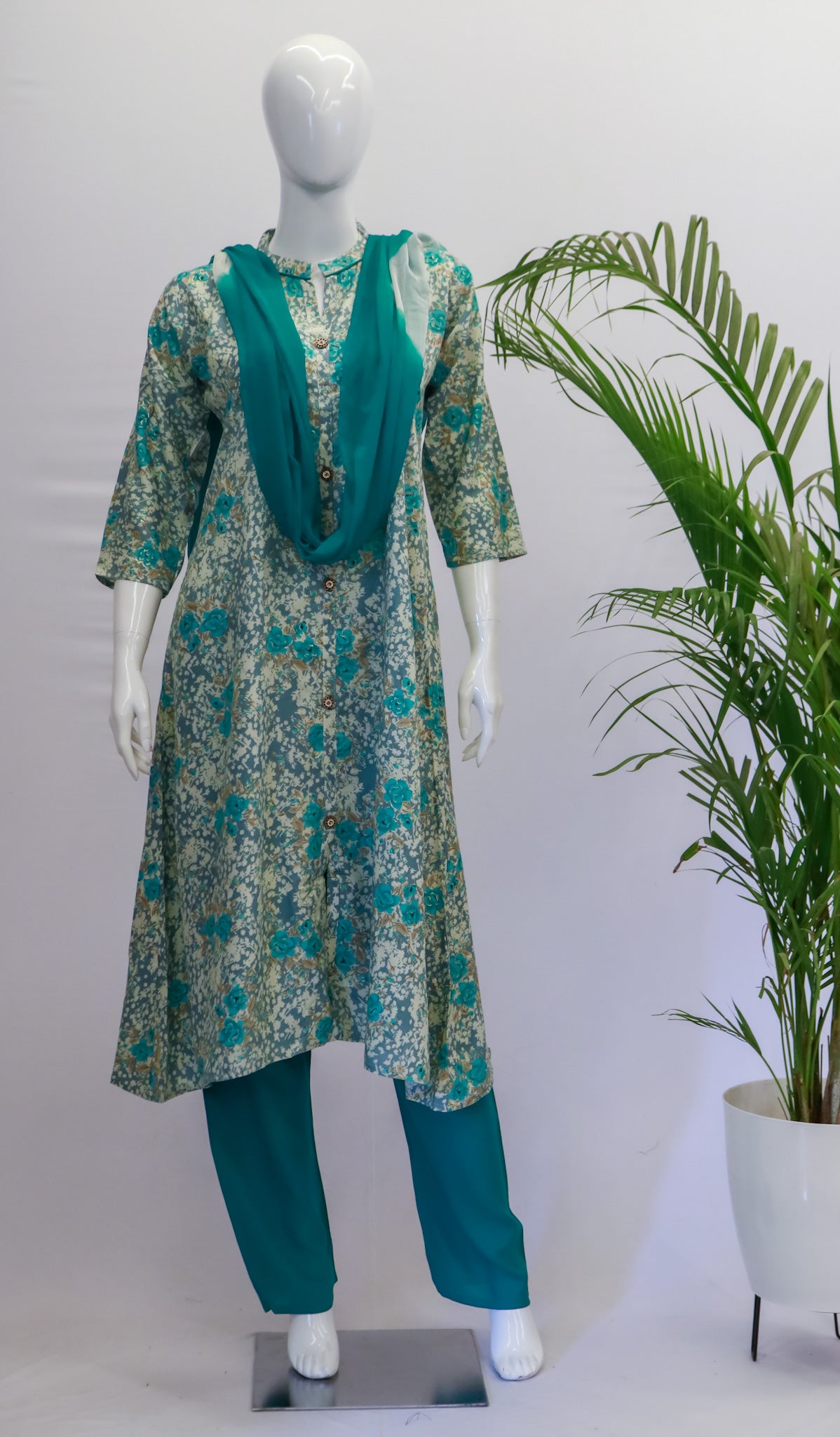 A mannequin displaying a blue dupatta set with floral print, consisting of a kurta, pant, and dupatta, standing in a retail setting with a potted plant in the background.
