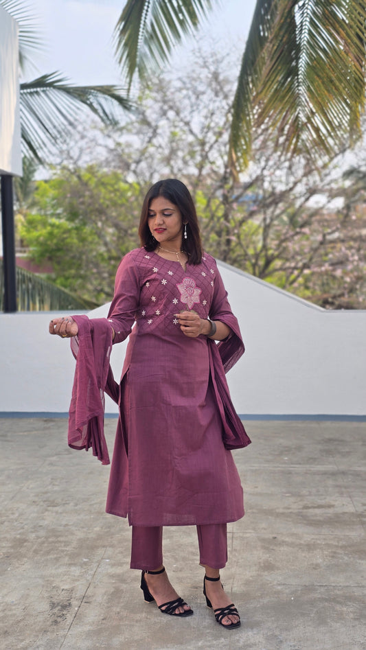 Woman in a purple outfit standing outdoors with trees and a building in the background