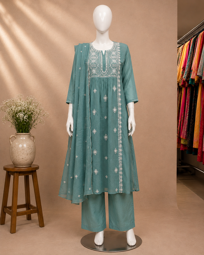 A teal green women's kurta set displayed on a white mannequin, featuring a long tunic with white embroidery, matching wide-leg trousers, and a coordinating dupatta. The set is photographed in a boutique setting with a beige wall, a wooden stool with a vase of flowers, and colorful garments hanging in the background.