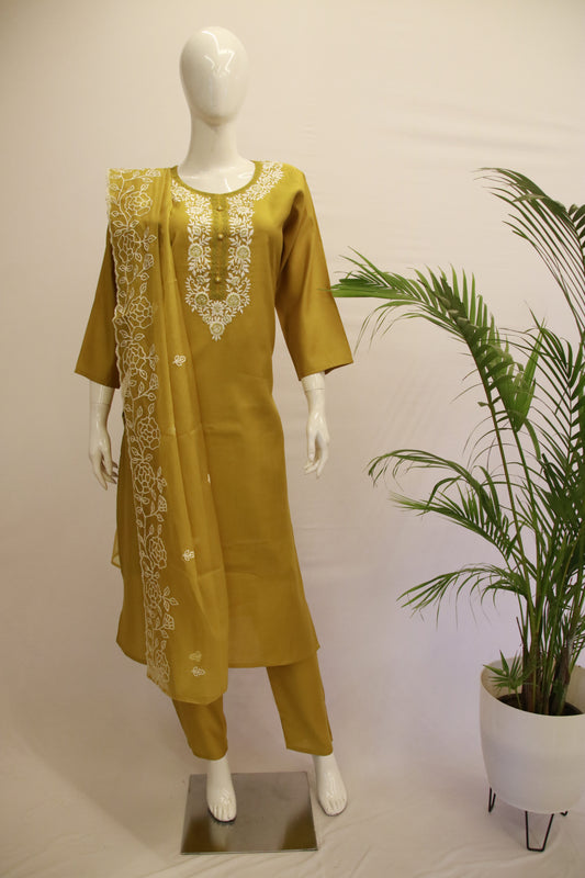 A mustard yellow silk chikankari dupatta set displayed on a mannequin, featuring a kurta with round-neck and chikankari work, and matching pants with elastic at the waist. There is a potted plant beside the mannequin.