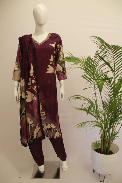 A mannequin displaying a purple dupatta set with floral prints, consisting of a kurta, pant, and dupatta, standing in a room with a potted plant behind it.
