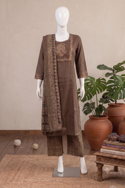 A brown three-piece salwar kameez suit displayed on a white mannequin, featuring a kurta with embroidery, matching trousers, and a dupatta with geometric borders, set against a neutral background with potted plants and a wooden table.